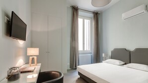 Ein modernes Hotelzimmer mit Bett, Schreibtisch mit Lampe, Fernseher und Blick durch ein Fenster mit Vorhängen.