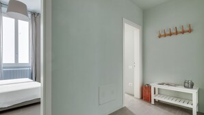 Ein Zimmer mit einem Bett, einer weißen Bank, einem Kleiderhaken und einem Fenster mit Vorhängen.