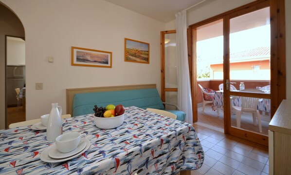 Une pièce comprenant un lit, une table avec un panier de fruits, une chaise, une table à manger avec des chaises, et donnant sur l’extérieur.