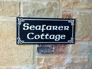 Ein schwarzes Schild mit weißer Schrift, das 'Seafarer Cottage' an einer Steinmauer zeigt.