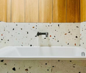 Een badkamer met een witte badkuip, een zwarte kraan en een spatwand van terrazzo tegels.