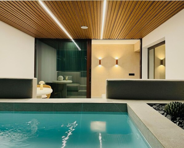 Un área de piscina interior moderna con techo de madera, dos zonas de estar y una puerta de vidrio que da a un baño.