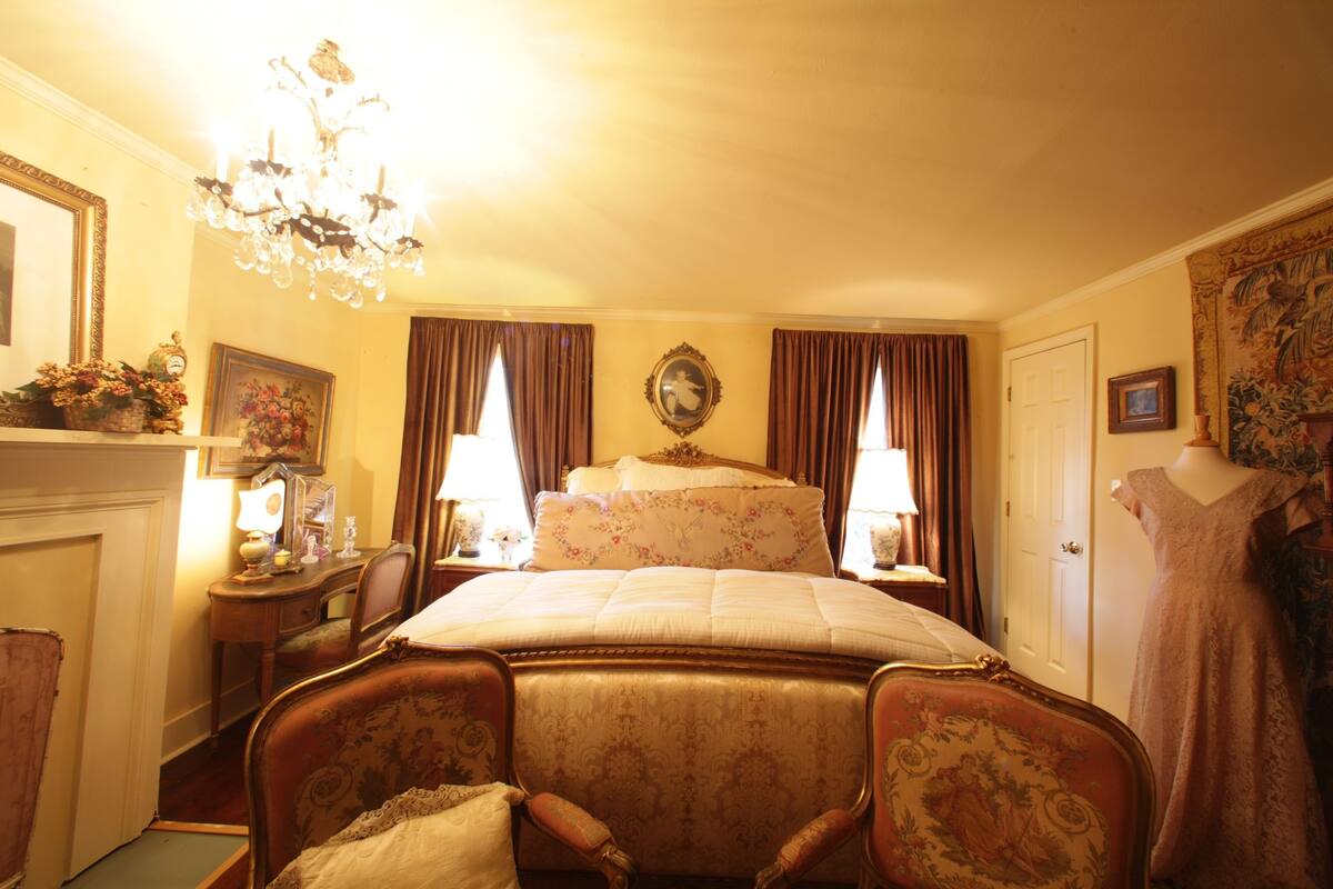 A classic bedroom with a large bed, a chandelier, a dresser, and a dress on a mannequin.