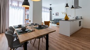 Un espace repas moderne avec une table en bois dressée pour six personnes, entourée de chaises grises. On aperçoit à l’arrière-plan une cuisine avec un îlot central blanc et un canapé gris.
