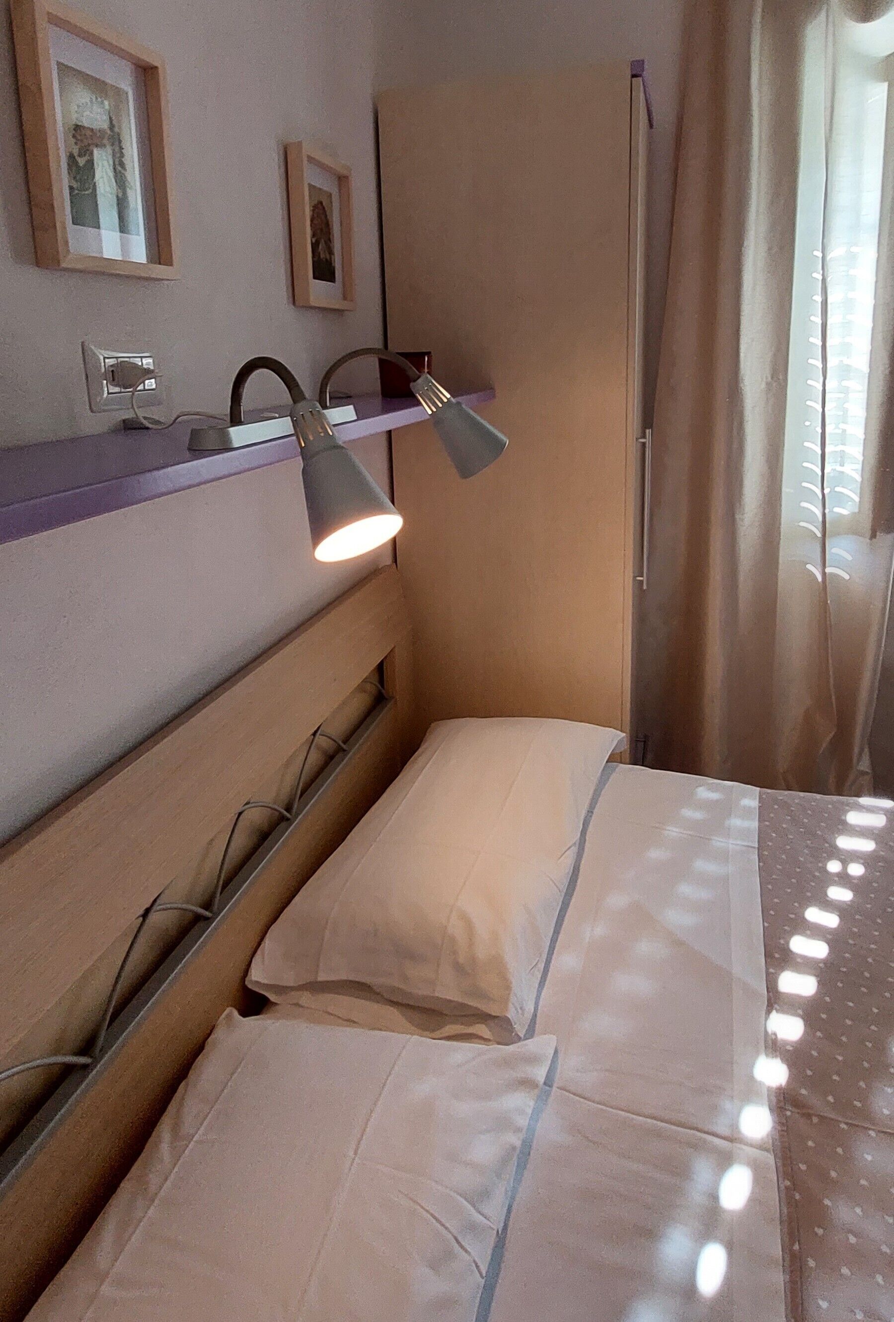 A hotel room with a bed, a headboard, a shelf with a lamp, and framed pictures on the wall.