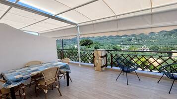 A balcony with a table and chairs, a view of a green landscape, and a white canopy.