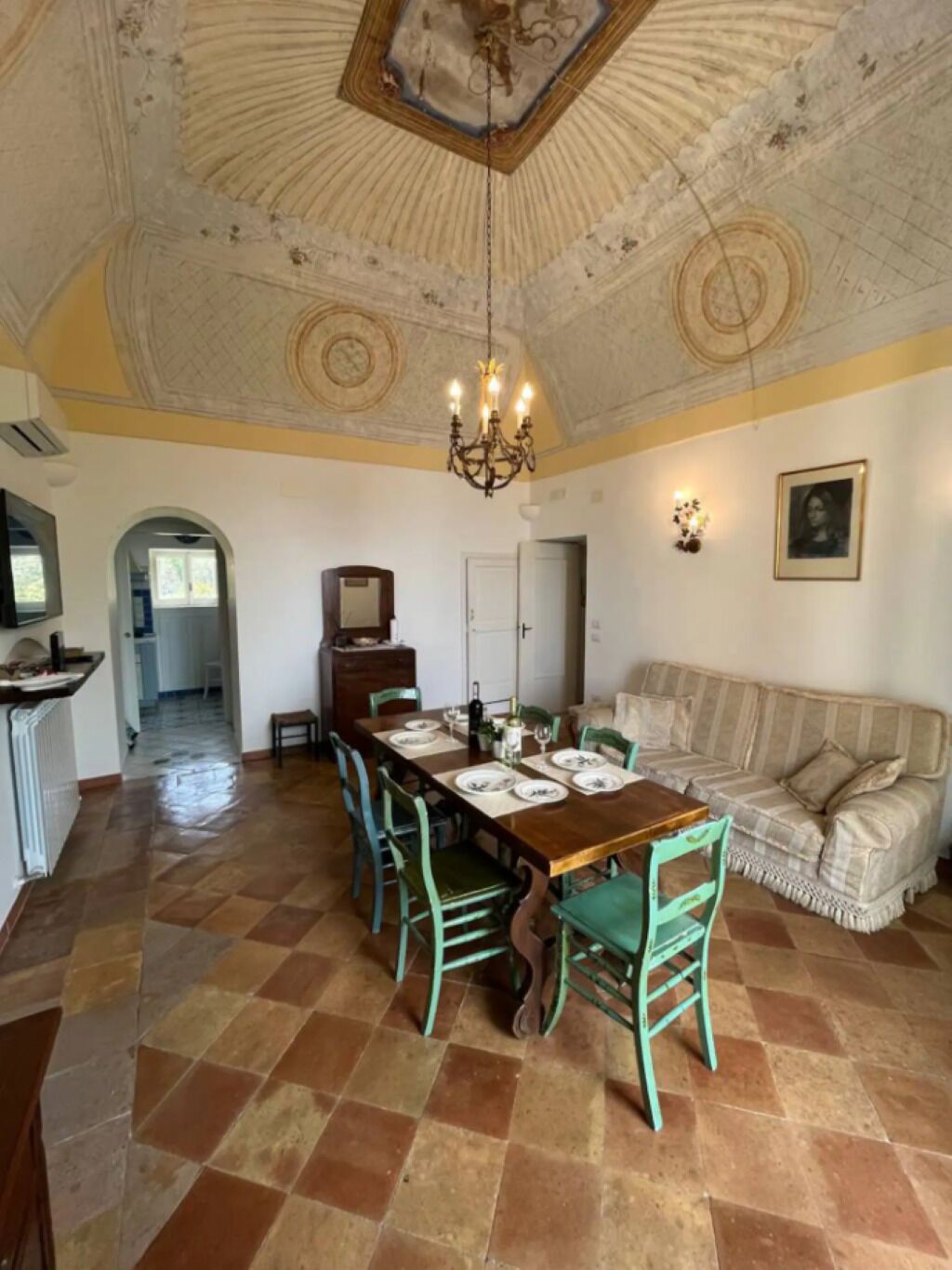 Ein Essbereich mit einem Holztisch, gedeckt für vier Personen, umgeben von grünen Stühlen. Eine Kristallleuchter hängt von der Decke, und ein Sofa ist im Hintergrund zu sehen.