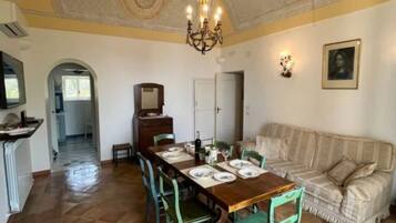 Ein Essbereich mit einem Holztisch, gedeckt für vier Personen, umgeben von grünen Stühlen. Eine Kristallleuchter hängt von der Decke, und ein Sofa ist im Hintergrund zu sehen.