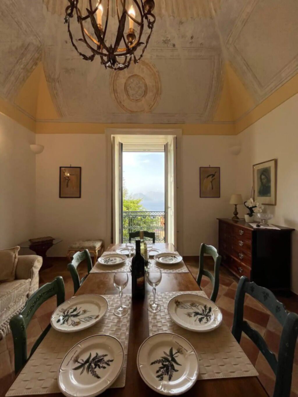 Ein Esszimmer mit einem langen Tisch, gedeckt für vier Personen, einer Chandelüster und Blick durch ein Fenster.
