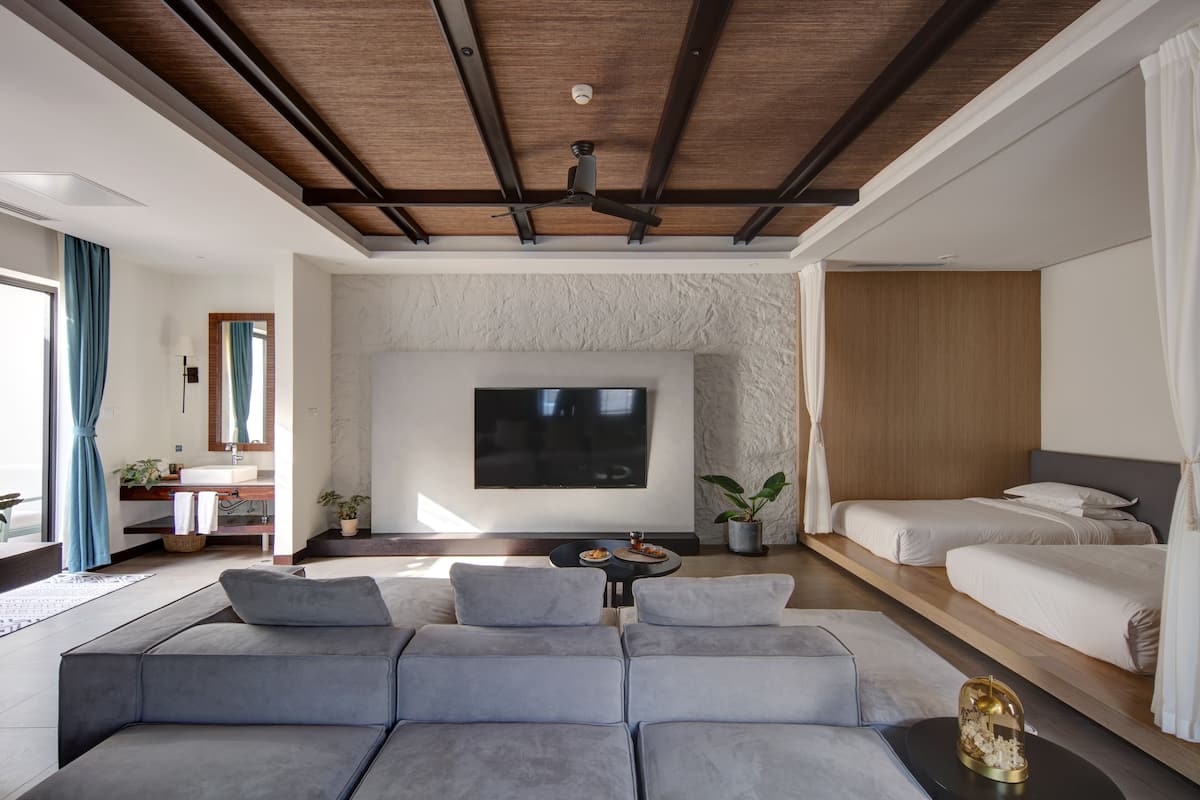 A modern hotel room with a large gray sofa, a flat-screen TV mounted on the wall, a wooden ceiling, and a bed with white bedding.