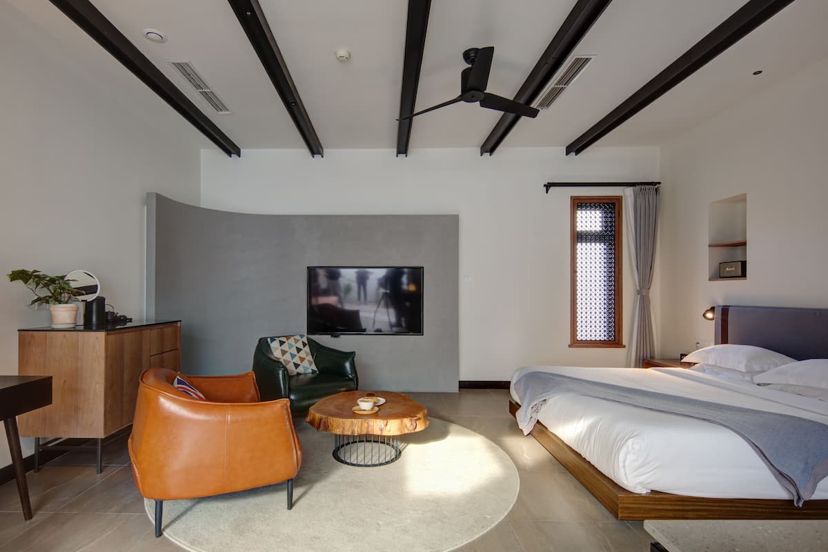 A modern bedroom with a bed, a TV mounted on the wall, a wooden desk, a leather chair, and a round coffee table.