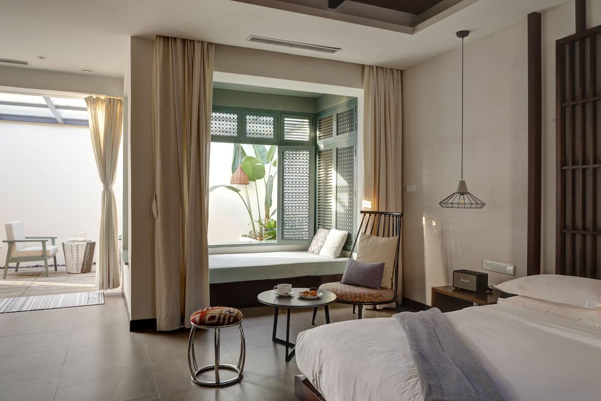 A hotel room with a bed, a chair, a small table with a cup and saucer, and a window with curtains.