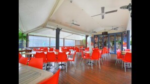 A covered dining area with red chairs and tables, ceiling fans, and large windows. - Gold Coast Family Apartment (Mermaid Beach)