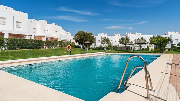 A swimming pool with a metal ladder, surrounded by a fence, and white buildings in the background.