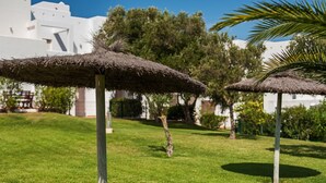 A well-maintained lawn with thatched umbrellas, a tree, and a white building in the background.