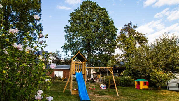 Una casita de juegos de madera con un tobogán azul, rodeada de árboles y un jardín cubierto de césped.