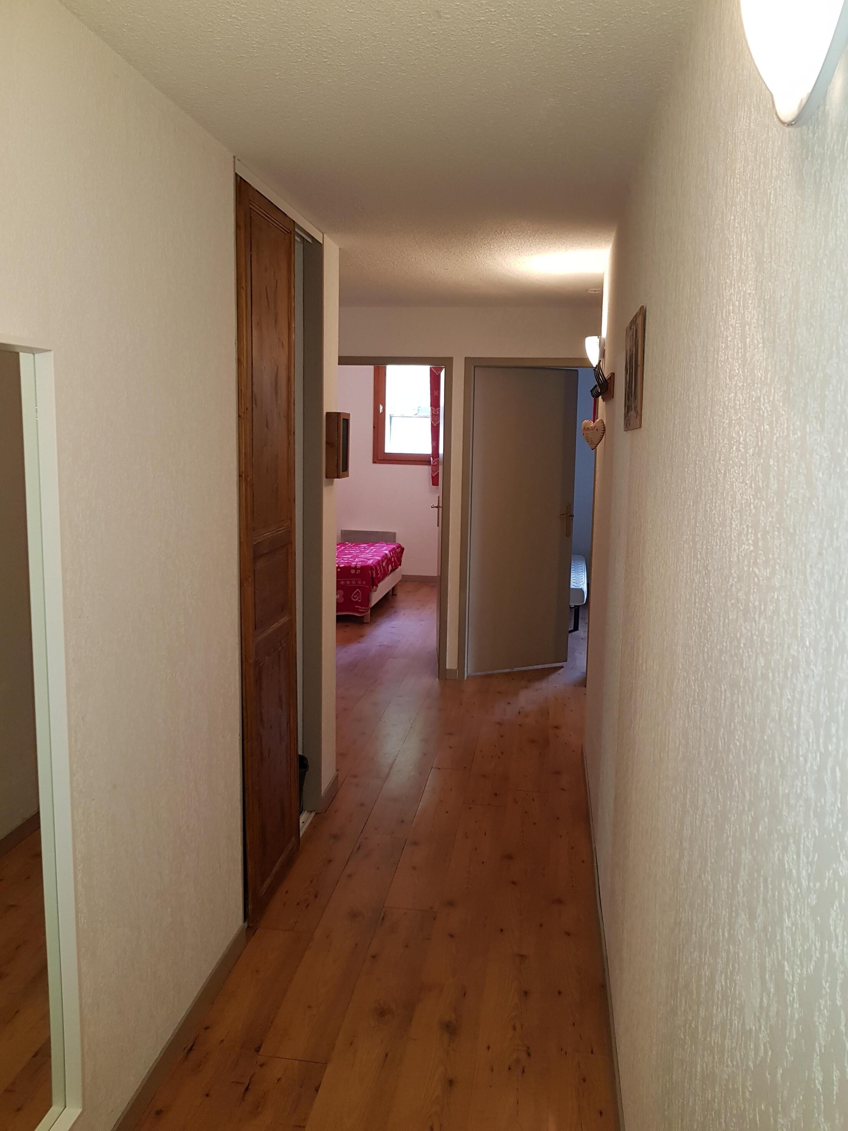 A narrow hallway with wooden flooring leading to a bedroom with a red bedspread.