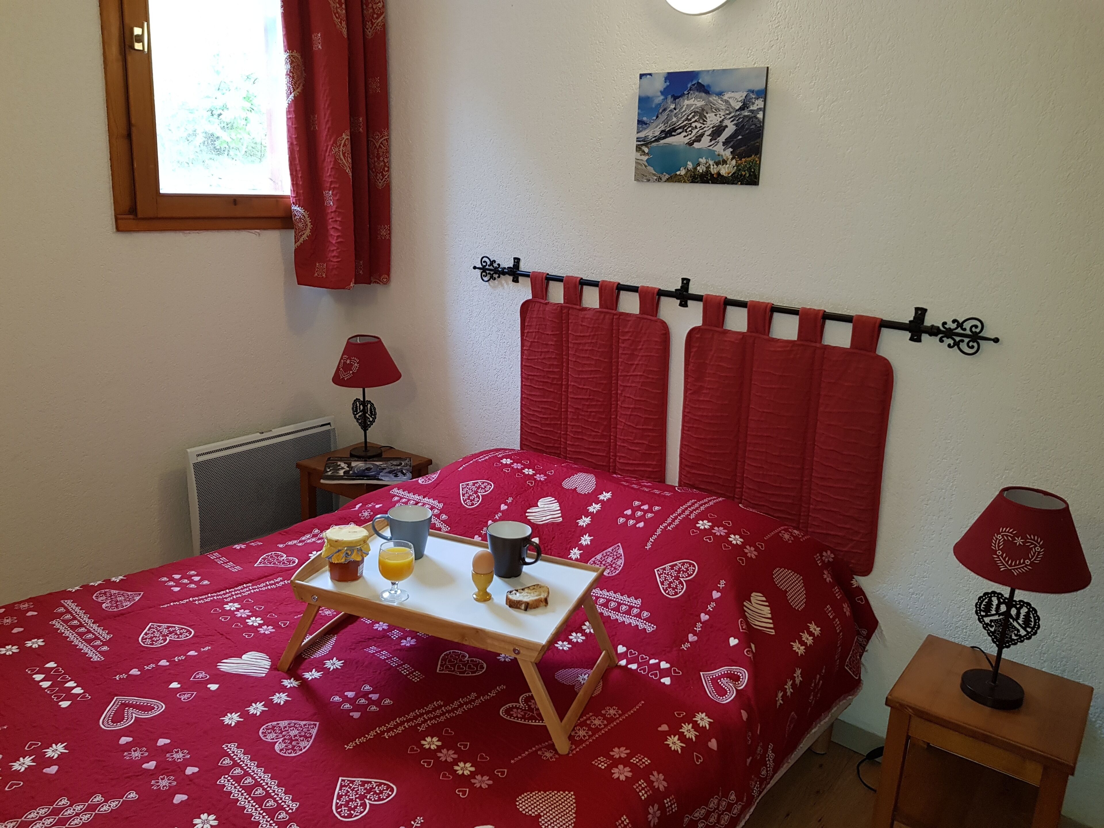 Ein Schlafzimmer mit roter Bettdecke, einem Nachttisch mit Lampe, einem Fenster mit Vorhängen und einem Bild an der Wand.