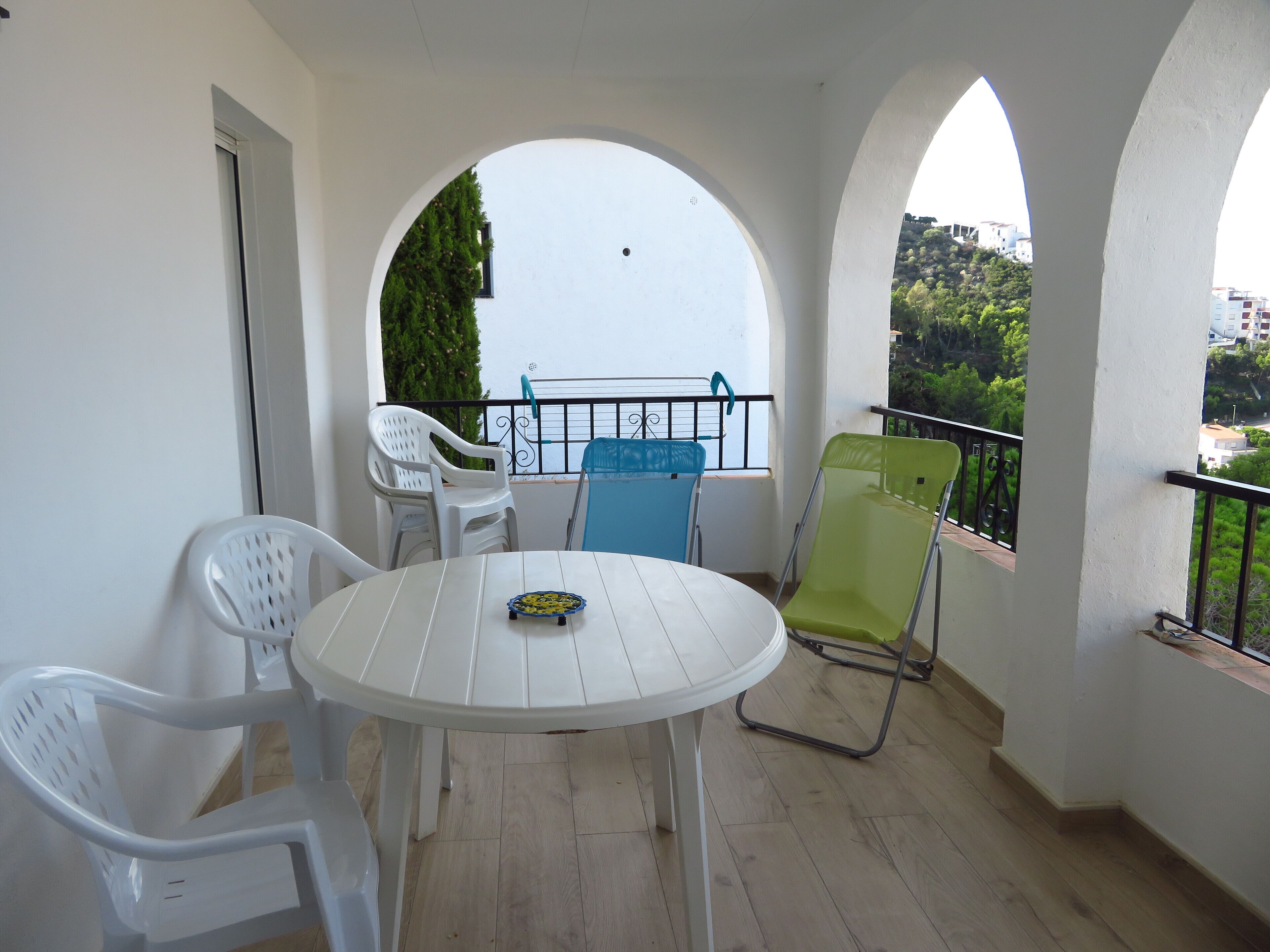 A balcony with a white table and chairs, a view of greenery and buildings, and a railing.