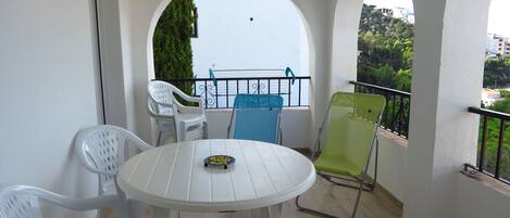 A balcony with a white table and chairs, a view of greenery and buildings, and a railing.