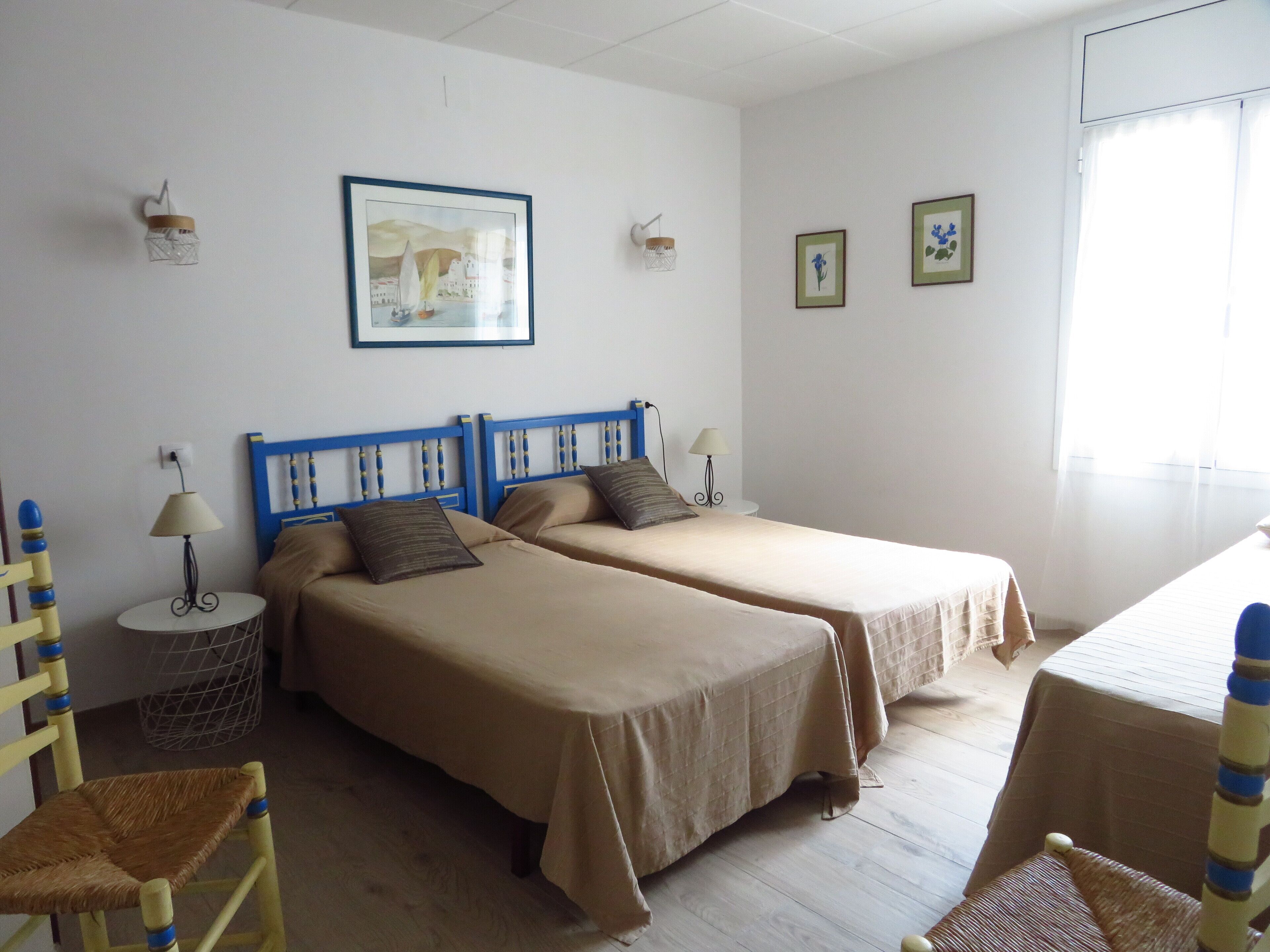 Una habitación con dos camas individuales, una mesita, una silla y una lámpara de pared.