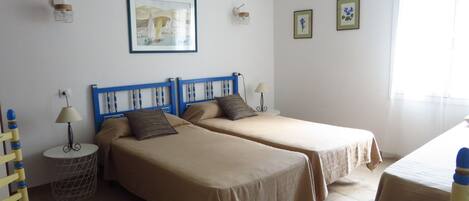 Una habitación con dos camas individuales, una mesita, una silla y una lámpara de pared.