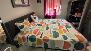 Ein Schlafzimmer mit Bett, Kopfteil, Wandbild, Fenster mit Vorhängen und Ablagefläche mit gefalteten Handtüchern.
