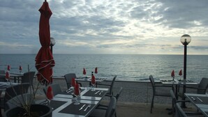 An outdoor dining area with tables and chairs, red umbrellas, and a view of the sea under a cloudy sky. - Studio sea view, wifi, spacious + balcony, near beach and shops (Menton)