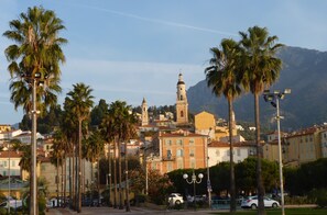 A coastal town with a prominent church tower, palm trees lining the streets, and a clear sky. - Studio sea view, wifi, spacious + balcony, near beach and shops (Menton)