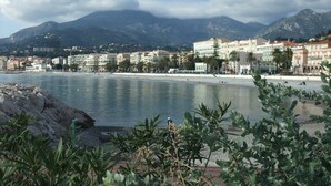 A coastal cityscape with a beach, buildings, and mountains in the background. - Studio sea view, wifi, spacious + balcony, near beach and shops (Menton)