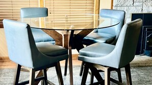 Une pièce avec une table basse dont le plateau est en verre, entourée de quatre chaises aux pieds en bois. On y trouve une cheminée en pierre, et le manteau de la cheminée est décoré de divers objets.