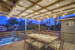 En uteplats med ett träbord dukat för middag, omgiven av stolar, under en pergola som är dekorerad med ljusslingor.