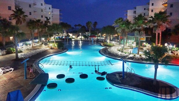 Una piscina con un área de juegos cercada por una red, rodeada de palmeras y sillas de descanso, con edificios al fondo.