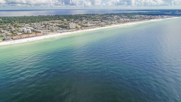 Vista aérea de una ciudad costera con una playa extensa, aguas azules cristalinas y una lejana vista del océano.