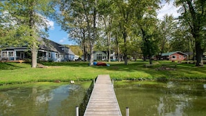 Une jetée en bois menant à une étendue d’eau calme, entourée d’une végétation luxuriante et de maisons.
