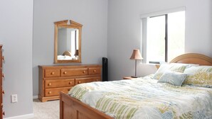 Une chambre à coucher avec une commode en bois, un lit avec une literie à motifs floraux, une table de chevet, une lampe et une fenêtre.