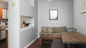 Un salon moderne avec un canapé, une table basse et une table à manger. Il y a un coin cuisine avec un évier et des placards. Une horloge est fixée au mur.