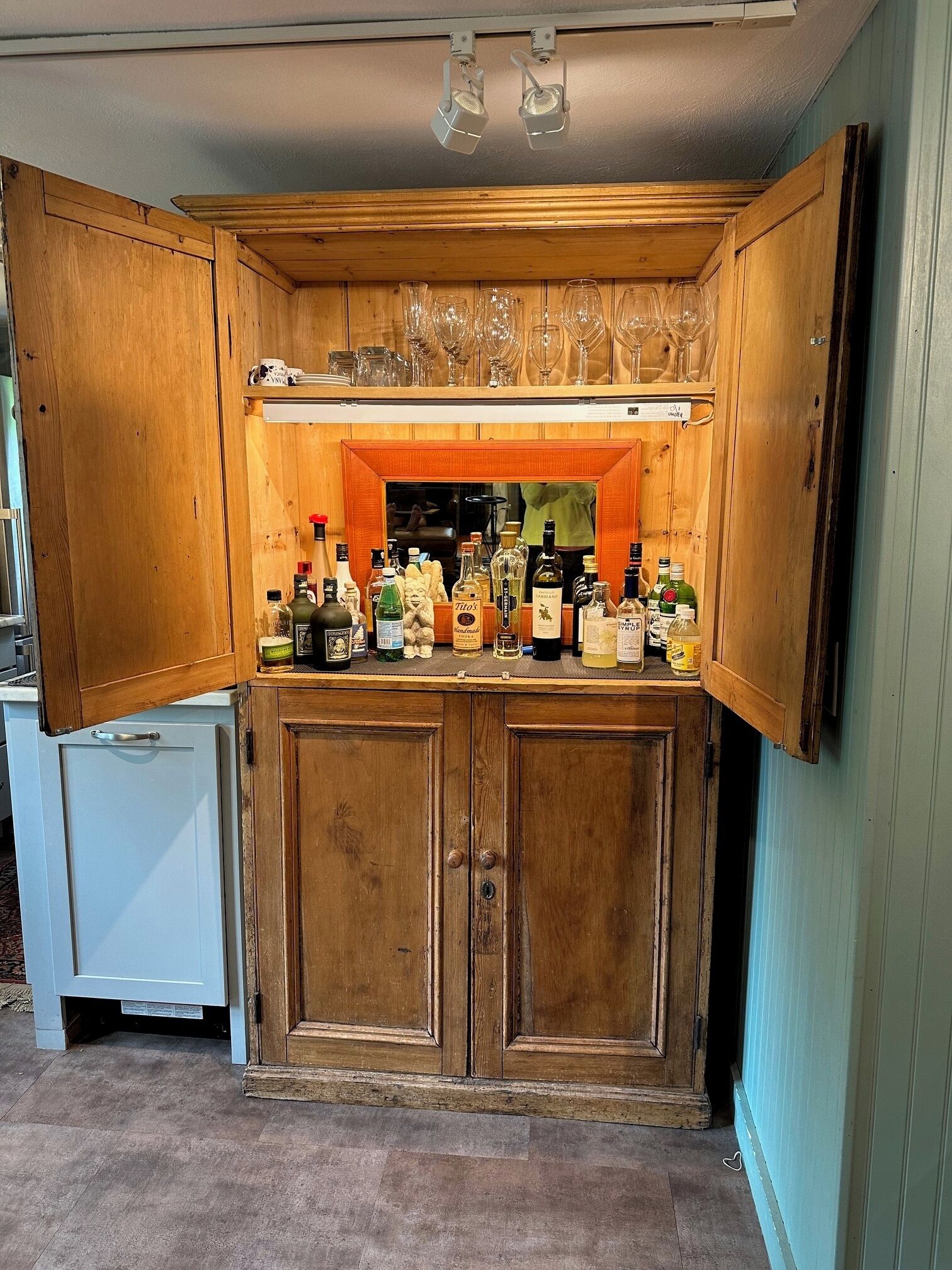 Un coin bar avec des meubles en bois, un comptoir sur lequel sont disposées différentes bouteilles, et une étagère au-dessus pour ranger les verres.