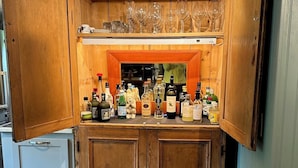A bar area with wooden cabinets, a countertop with various bottles, and a shelf above holding glasses.
