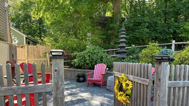 A wooden gate with a stone pathway leading to a garden area with red chairs and a lantern post.