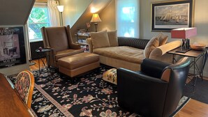 A cozy living room with a brown leather armchair, a beige sofa, and a black leather chair. There is a patterned rug on the floor, a small table with a lamp, and a framed picture on the wall.