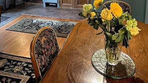 A dining area with a wooden table, chairs, and a vase of flowers. There is a kitchen in the background with cabinets, a refrigerator, and a microwave.