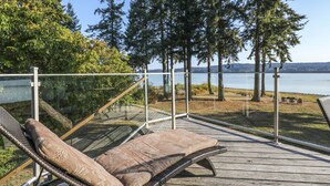 A wooden deck with lounge chairs and a glass railing overlooking a lake, surrounded by trees.