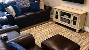 Un salon avec un canapé en cuir marron foncé, un pouf assorti, un meuble en bois clair et un téléviseur à écran plat fixé au mur.