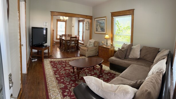 A living room with a sofa, a coffee table, a TV on a stand, a dining area with a table and chairs, and a window with blinds. - 1st Class Rentals - Munson Manor III at Murdock (Oneonta)