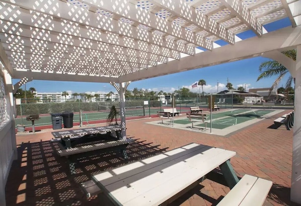 An outdoor tennis court area with a covered seating area, benches, and a grill.