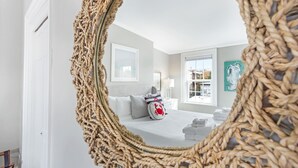 Ein Schlafzimmer mit einem großen gewebten Spiegel, einem Bett mit weißer Bettwäsche, einem Nachttisch, einer Lampe und Blick ins Freie durch ein Fenster.