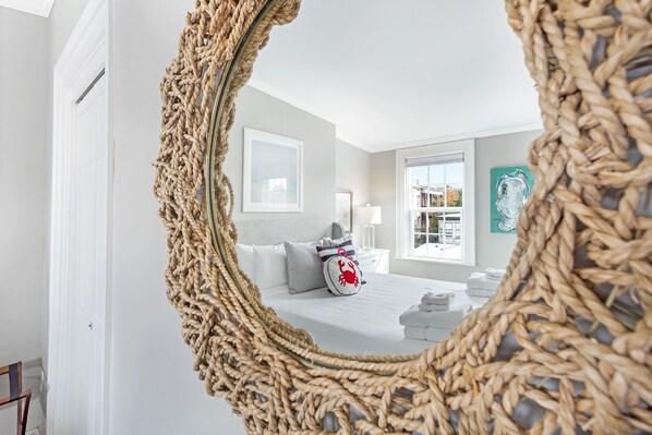 A bedroom with a large woven mirror, a bed with white linens, a nightstand, a lamp, and a view of the outdoors through a window.