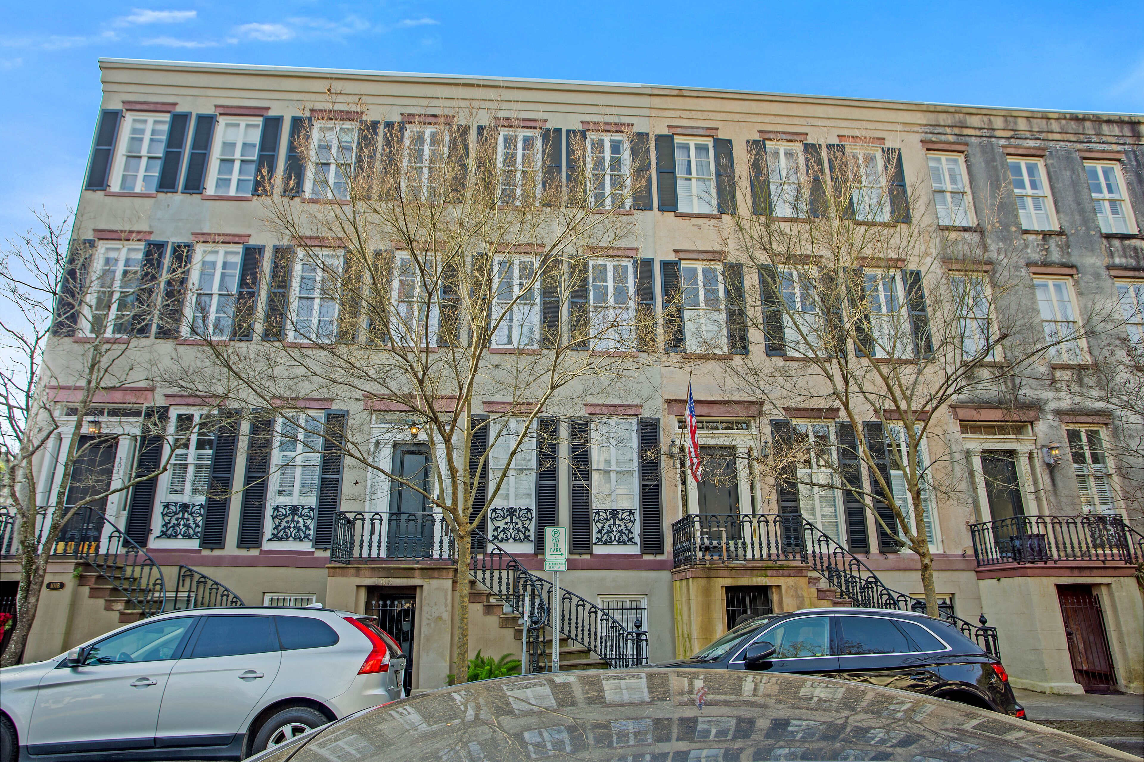 A multi-story building with a row of windows and balconies, a staircase leading to the entrance, and parked cars in front.