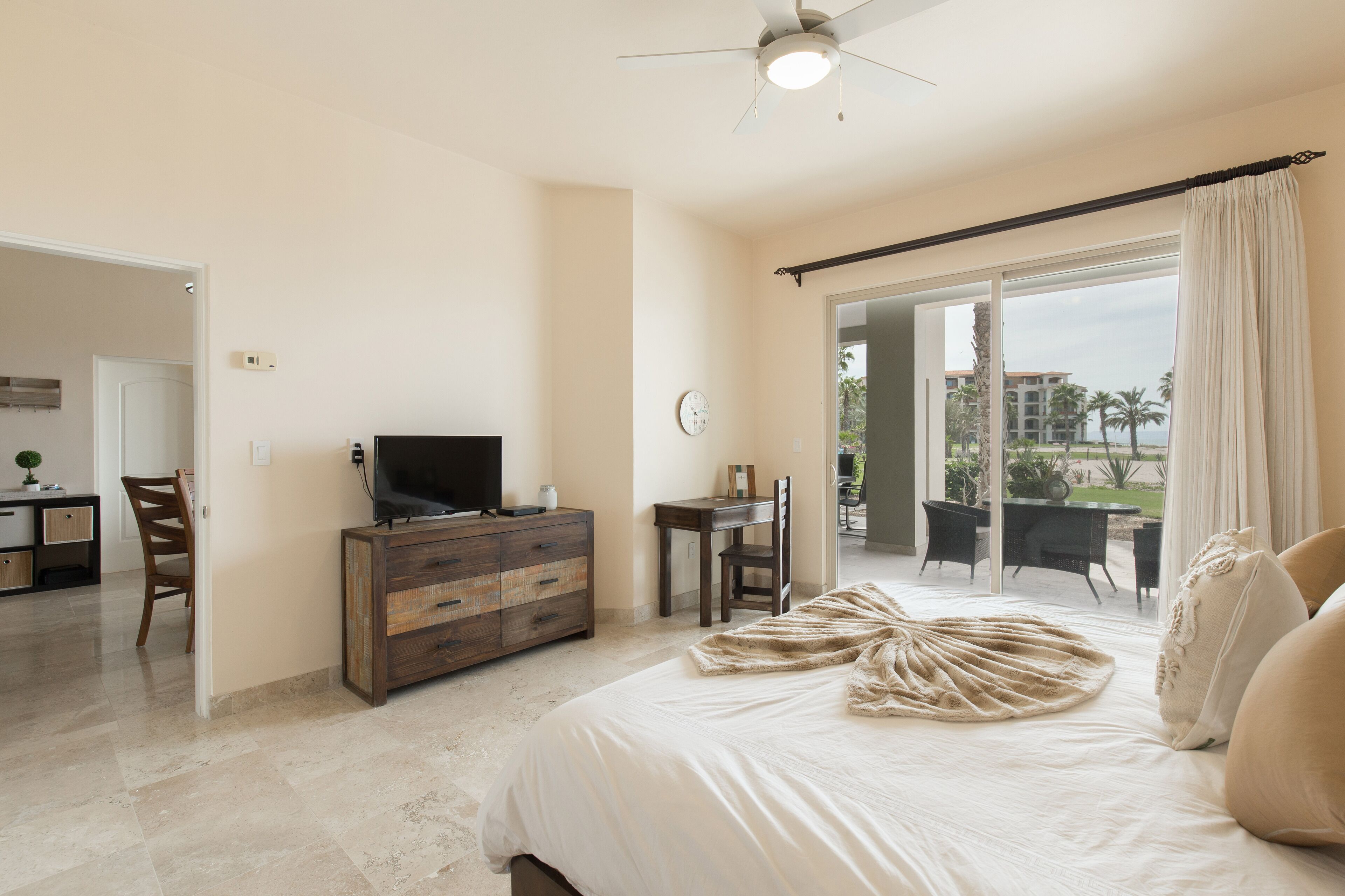 A bedroom with a bed, a TV, a desk, and a chair. There is a balcony with a view of palm trees and buildings.